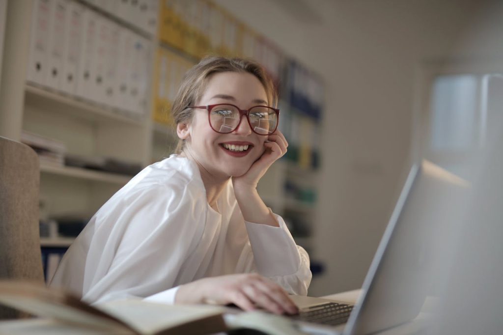 A cheerful woman with glasses working on a laptop in an office environment, showing modern technology use.